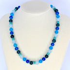A beaded necklace with a gradient of blue hues, from light sky blue to deep navy. The necklace features round beads interspersed with smaller, cylindrical beads, creating a visually appealing accessory. The necklace is displayed on a white mannequin.