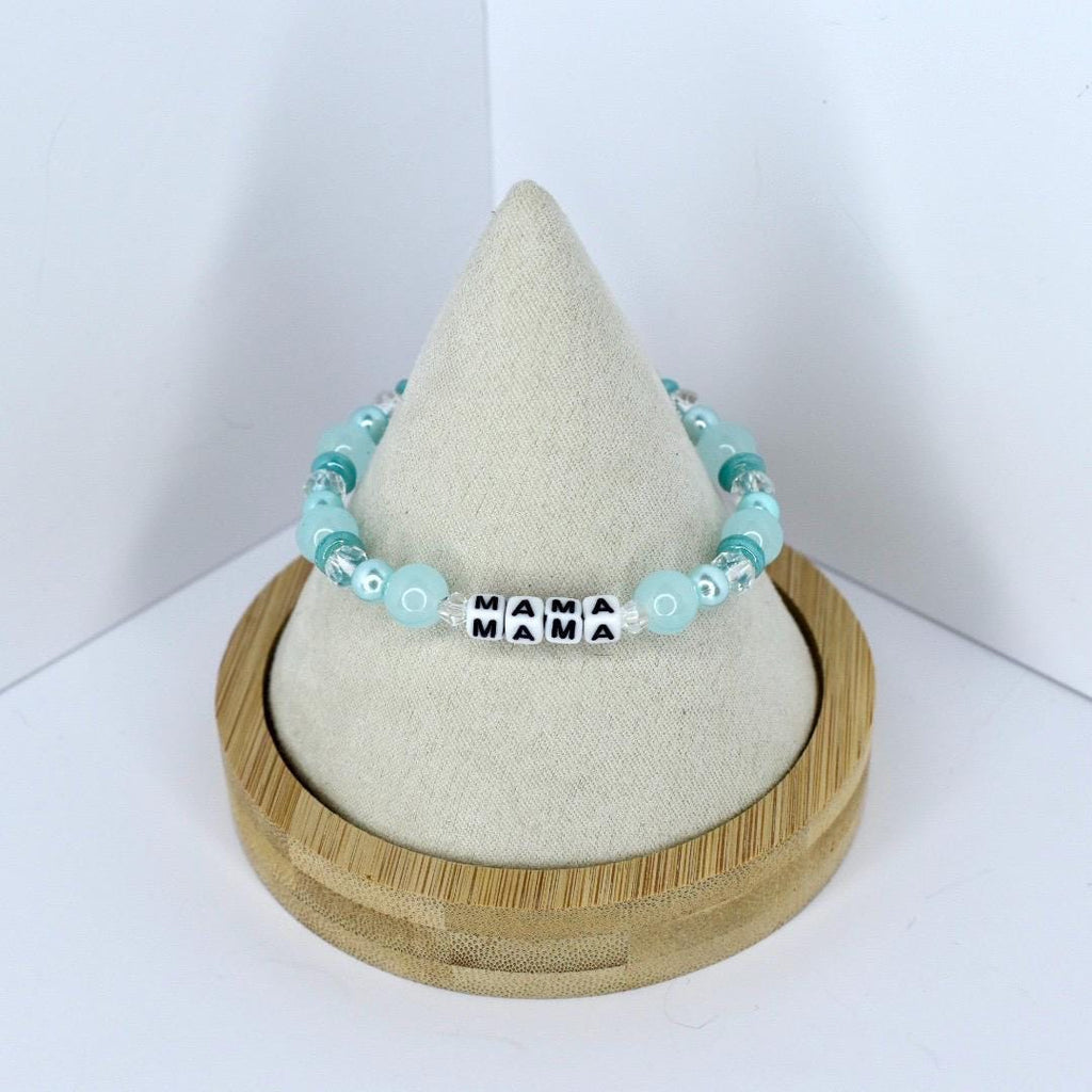 A beaded bracelet with the word "MAMA" spelled out in black and white letter beads. The bracelet features light blue and clear beads. The bracelet is displayed on a white cone-shaped stand inside a wooden holder.