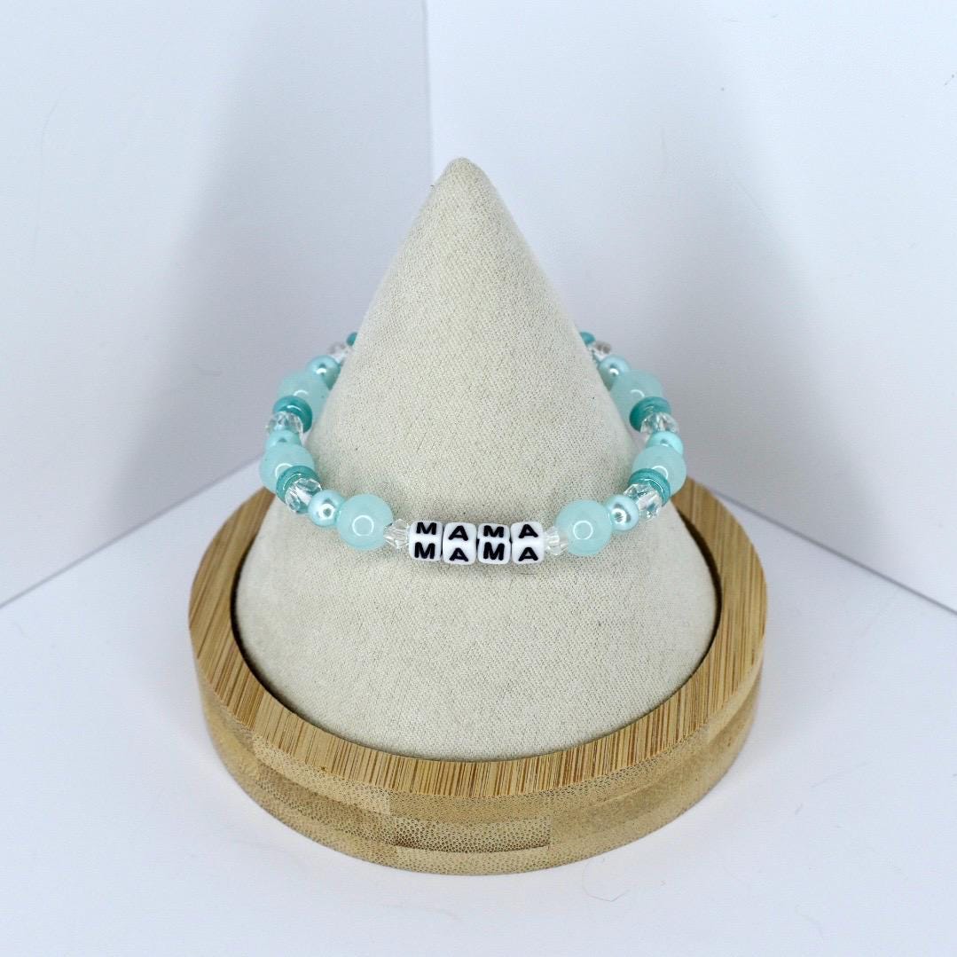 A beaded bracelet with the word "MAMA" spelled out in black and white letter beads. The bracelet features light blue and clear beads. The bracelet is displayed on a white cone-shaped stand inside a wooden holder.