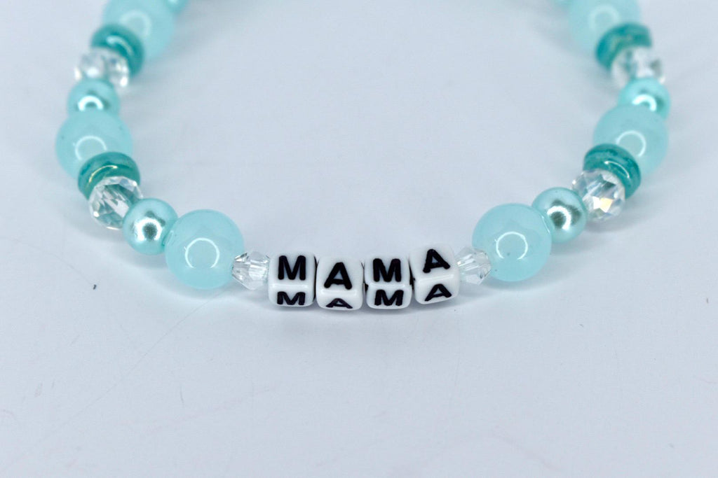 A beaded bracelet with the word "MAMA" spelled out in black letters on white square beads. The bracelet features light blue and teal beads, with clear crystal accents. The bracelet is on a white background.