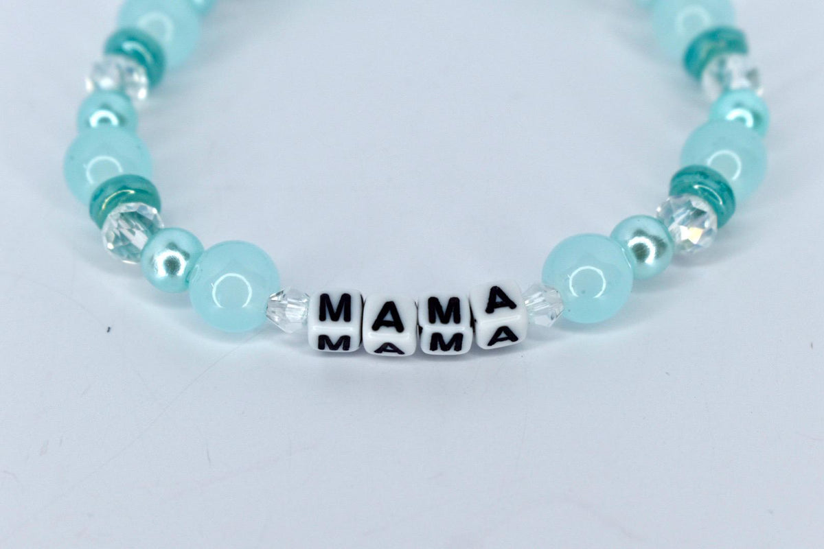 A beaded bracelet with the word "MAMA" spelled out in black letters on white square beads. The bracelet features light blue and teal beads, with clear crystal accents. The bracelet is on a white background.