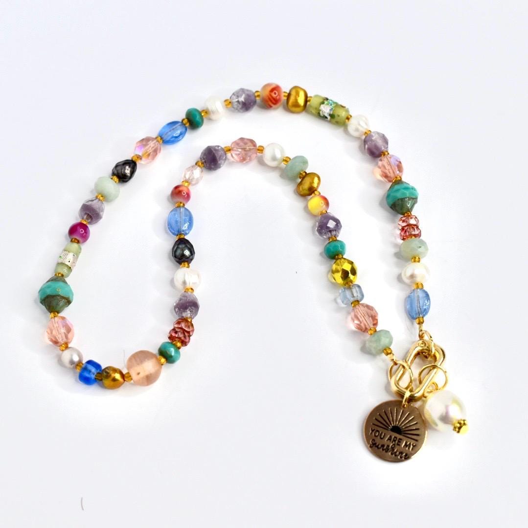 A colorful beaded necklace with a variety of shapes and sizes of beads in shades of pink, blue, green, yellow, and gold. A gold-colored charm with the words "YOU ARE MY sunshine" hangs from the necklace, along with a white pearl.