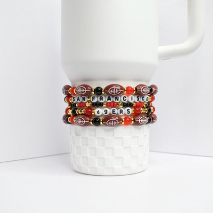 Four beaded bracelets stacked on a white ceramic mug. The bracelets feature a combination of red, black, and gold beads. Some beads are shaped like footballs, and others spell out 'SAN FRANCISCO 49ERS'.