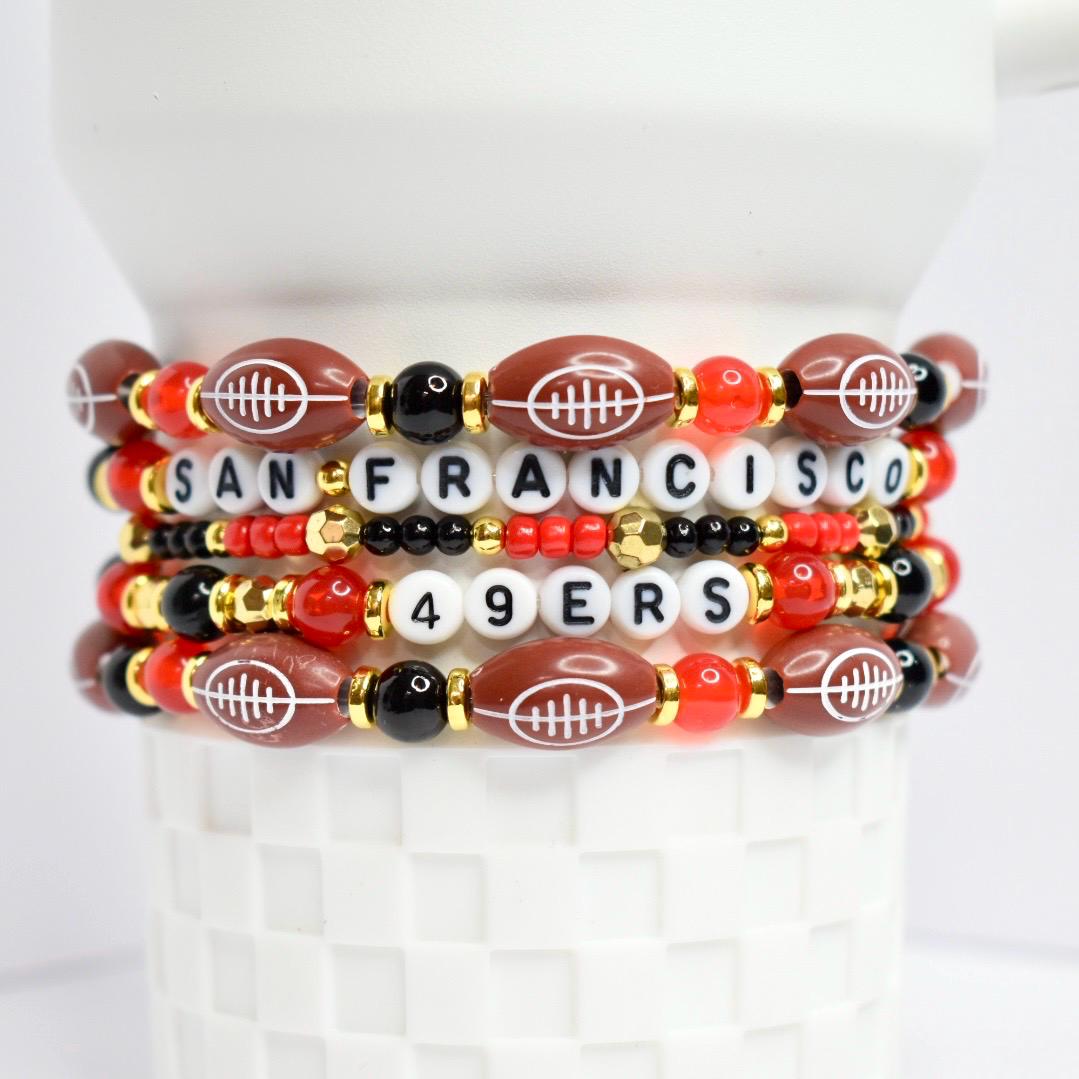 Stack of beaded bracelets featuring football-shaped beads and letter beads spelling 'SAN FRANCISCO' and '49ERS'. The bracelets have a color scheme of brown, red, black, gold, and white. The football beads have white stitching.