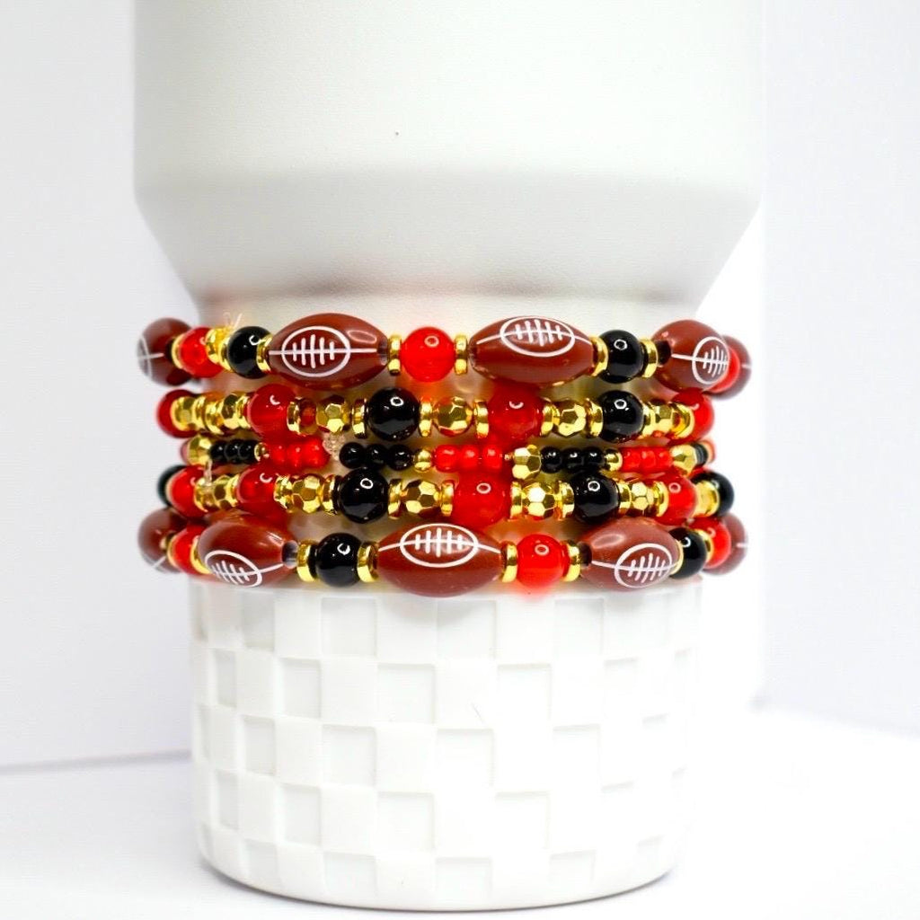 A stack of beaded bracelets featuring football-shaped beads. The bracelets alternate between brown football beads with white stitching, black beads, red beads, and gold-colored beads. The bracelets are stacked on a white, textured surface.