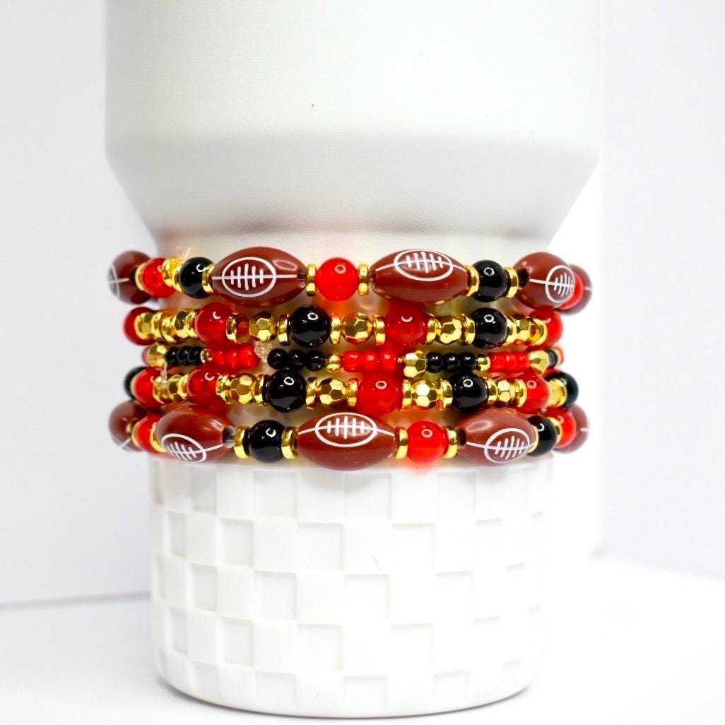 A stack of beaded bracelets featuring football-shaped beads. The bracelets alternate between brown football beads with white stitching, black beads, red beads, and gold-colored beads. The bracelets are stacked on a white, textured surface.