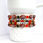 A stack of beaded bracelets featuring a football theme. The bracelets are primarily brown, red, black, and gold. Each bracelet has football-shaped beads, letter beads spelling 'SAN FRANCISCO 49ERS', and various round beads. The bracelets are stacked on a white, textured surface.