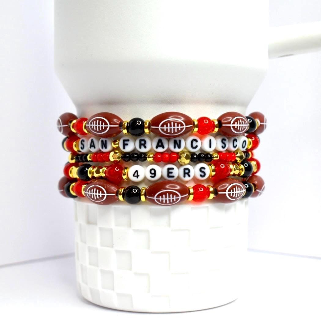 A stack of beaded bracelets featuring a football theme. The bracelets are primarily brown, red, black, and gold. Each bracelet has football-shaped beads, letter beads spelling 'SAN FRANCISCO 49ERS', and various round beads. The bracelets are stacked on a white, textured surface.