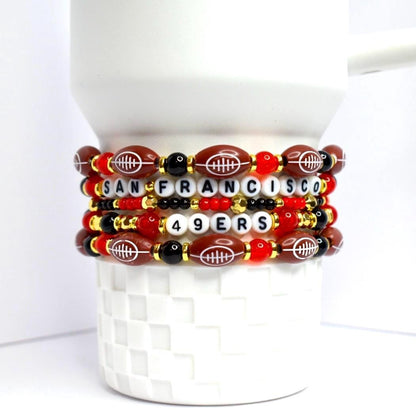 A stack of beaded bracelets featuring a football theme. The bracelets are primarily brown, red, black, and gold. Each bracelet has football-shaped beads, letter beads spelling 'SAN FRANCISCO 49ERS', and various round beads. The bracelets are stacked on a white, textured surface.