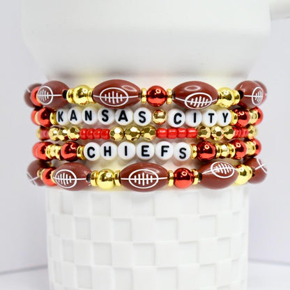 A stack of beaded bracelets featuring a Kansas City Chiefs theme. The bracelets are composed of football-shaped beads, white letter beads spelling 'KANSAS CITY CHIEFS', and gold and red accent beads. The bracelets are displayed on a white, textured surface.