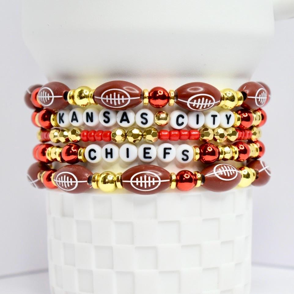 A stack of beaded bracelets featuring a Kansas City Chiefs theme. The bracelets are composed of football-shaped beads, white letter beads spelling 'KANSAS CITY CHIEFS', and gold and red accent beads. The bracelets are displayed on a white, textured surface.