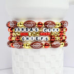 A stack of beaded bracelets featuring a Kansas City Chiefs theme. The bracelets are composed of football-shaped beads, white letter beads spelling 'KANSAS CITY CHIEFS', and gold and red accent beads. The bracelets are displayed on a white, textured surface.