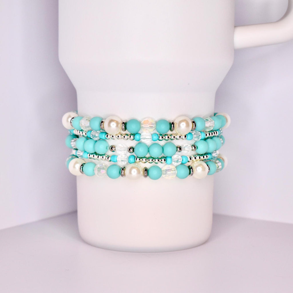 A close-up of a white tumbler with a handle, wrapped with five stacked beaded bracelets in a coordinated turquoise and white theme. The bracelets feature matte turquoise beads, iridescent clear beads, pearl-like white beads, and shiny silver spacers.