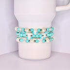 A close-up of a white tumbler with a handle, wrapped with five stacked beaded bracelets in a coordinated turquoise and white theme. The bracelets feature matte turquoise beads, iridescent clear beads, pearl-like white beads, and shiny silver spacers.
