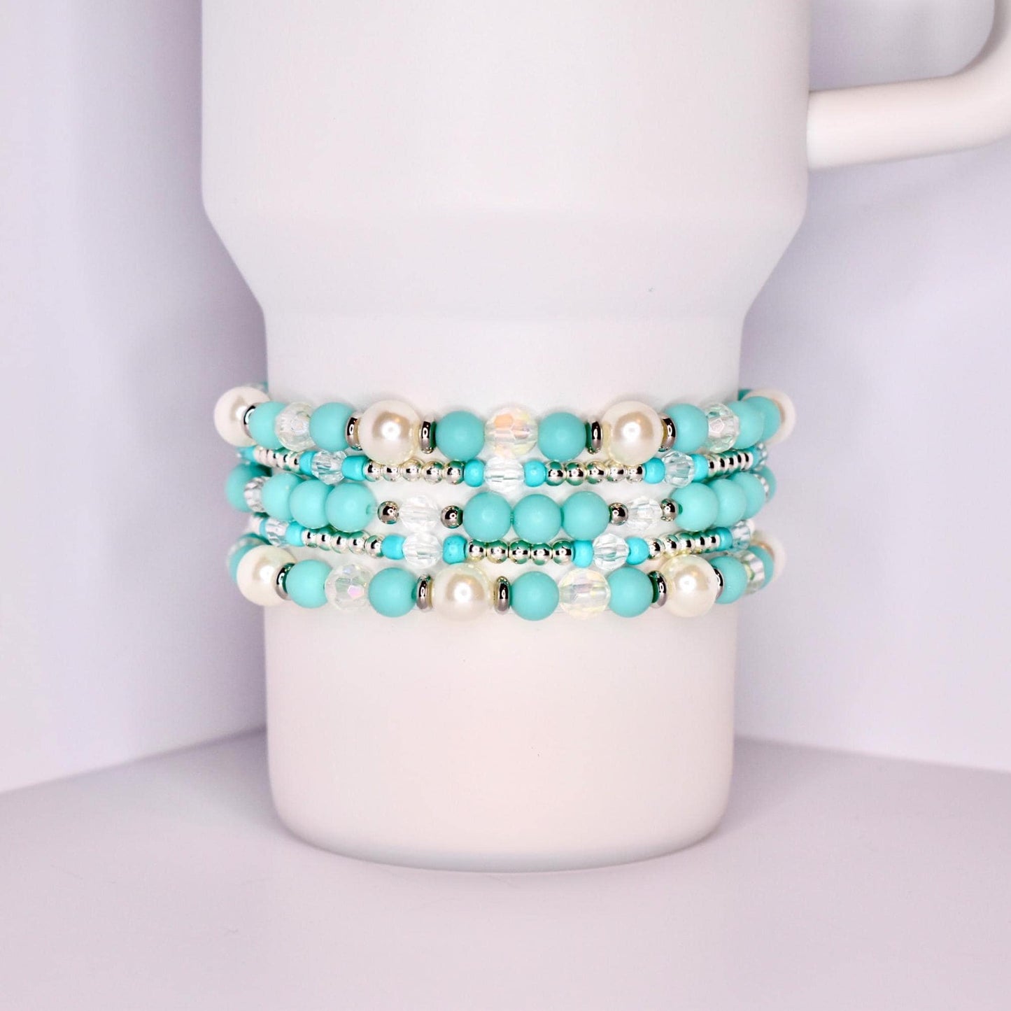 A close-up of a white tumbler with a handle, wrapped with five stacked beaded bracelets in a coordinated turquoise and white theme. The bracelets feature matte turquoise beads, iridescent clear beads, pearl-like white beads, and shiny silver spacers.