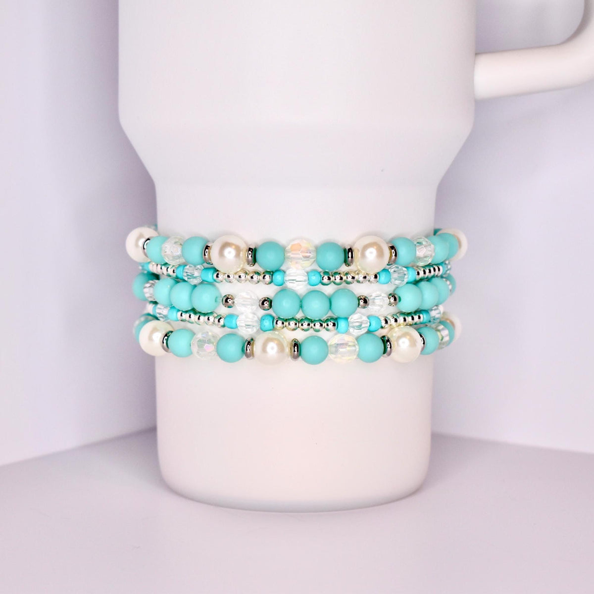 A close-up of a white tumbler with a handle, wrapped with five stacked beaded bracelets in a coordinated turquoise and white theme. The bracelets feature matte turquoise beads, iridescent clear beads, pearl-like white beads, and shiny silver spacers.