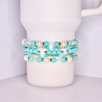 A close-up of a white tumbler with a handle, wrapped with five stacked beaded bracelets in a coordinated turquoise and white theme. The bracelets feature matte turquoise beads, iridescent clear beads, pearl-like white beads, and shiny silver spacers.