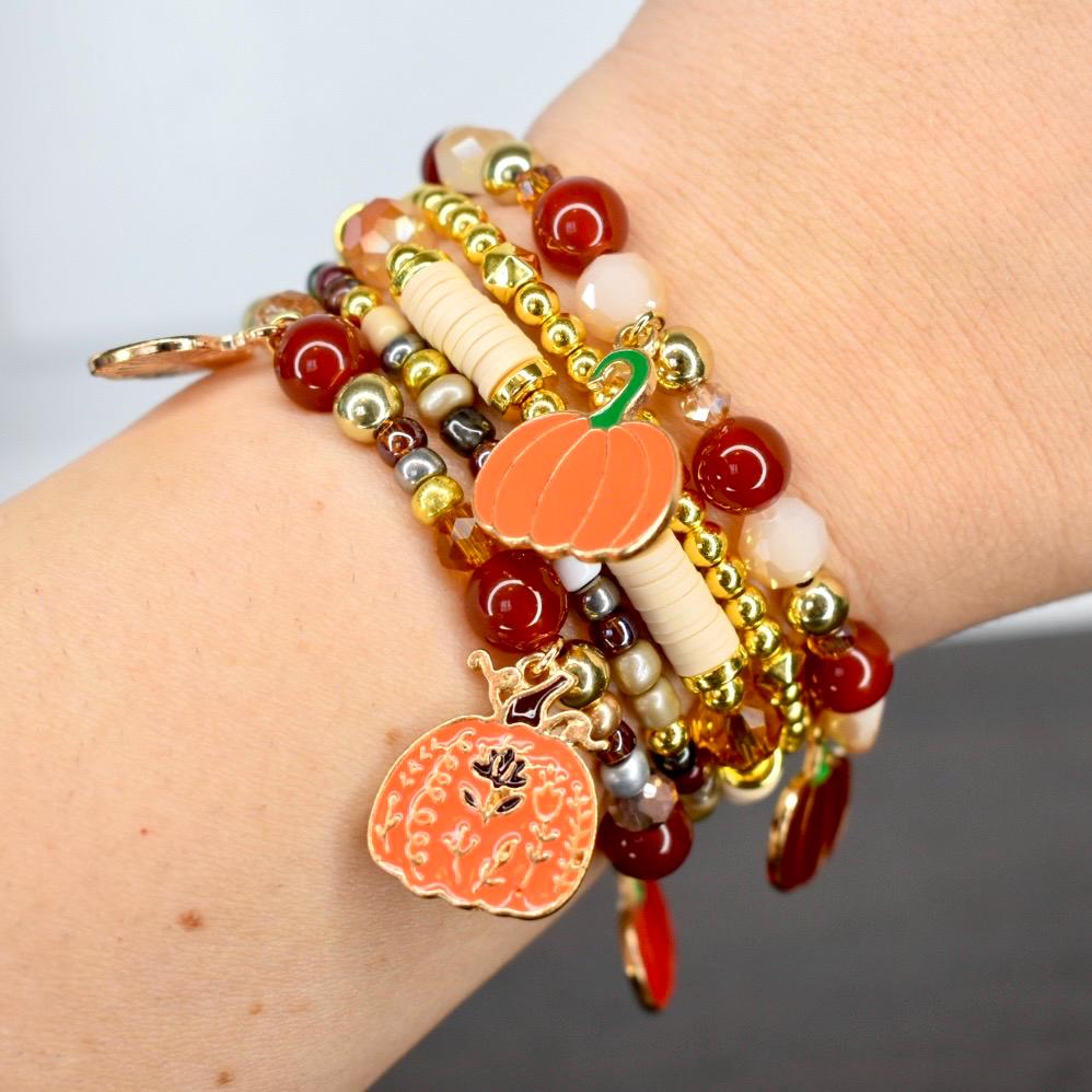 Autumn charm bracelet set on someone's wrist with pumpkin and harvest charms and dark red, gold, and neutral beads.