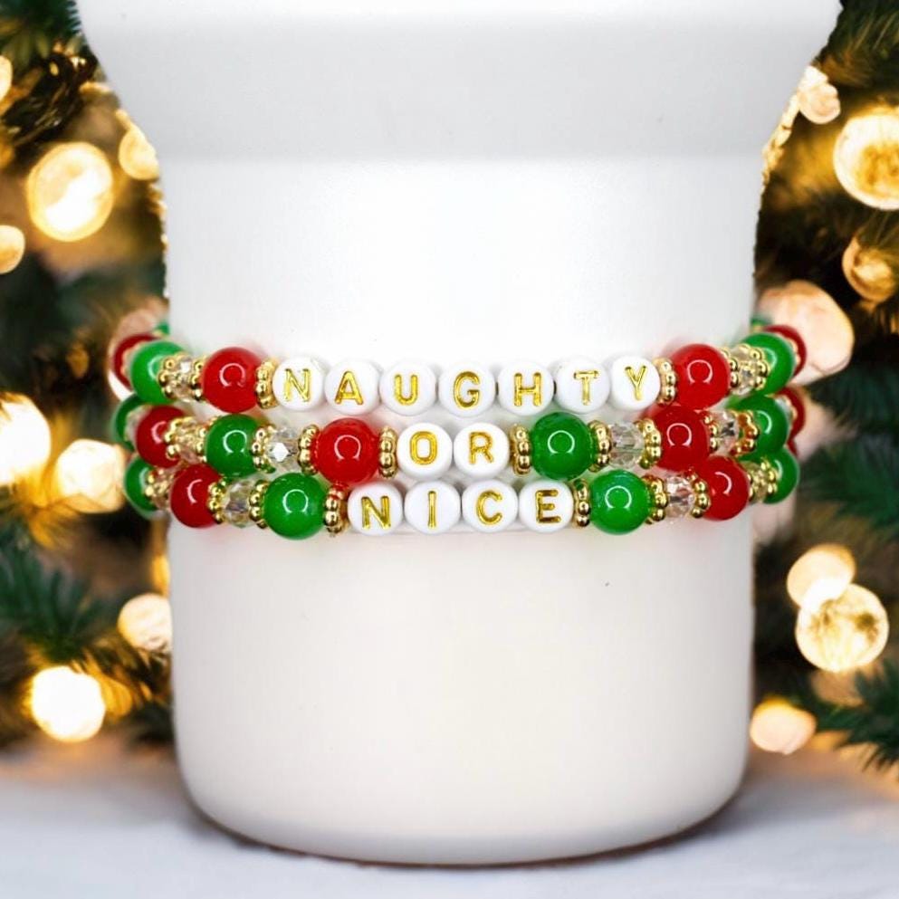 Three Christmas beaded bracelets with red, green, and white beads spelling out Naughty or Nice in gold letters, styled with holiday lights.