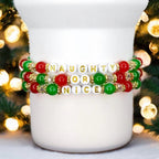 Three Christmas beaded bracelets with red, green, and white beads spelling out Naughty or Nice in gold letters, styled with holiday lights.