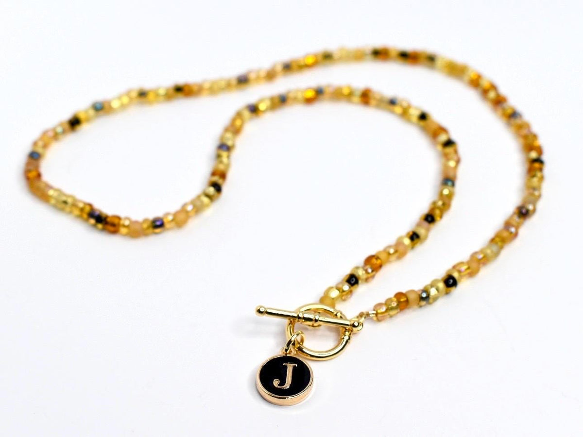 A beaded necklace featuring a toggle clasp and a circular pendant. The necklace is composed of small golden-yellow beads with interspersed darker brown and black beads. The pendant is black with a gold border and a white letter 'J' in the center. The toggle clasp and pendant's metal components are gold-colored.