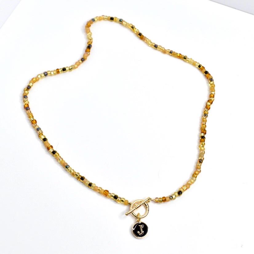 A beaded necklace with a gold toggle clasp and a black pendant. The necklace features small, round beads in shades of gold, brown, and black. The pendant is black with a gold letter 'J'. The necklace is displayed on a white surface.