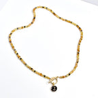 A beaded necklace with a gold toggle clasp and a black pendant. The necklace features small, round beads in shades of gold, brown, and black. The pendant is black with a gold letter 'J'. The necklace is displayed on a white surface.