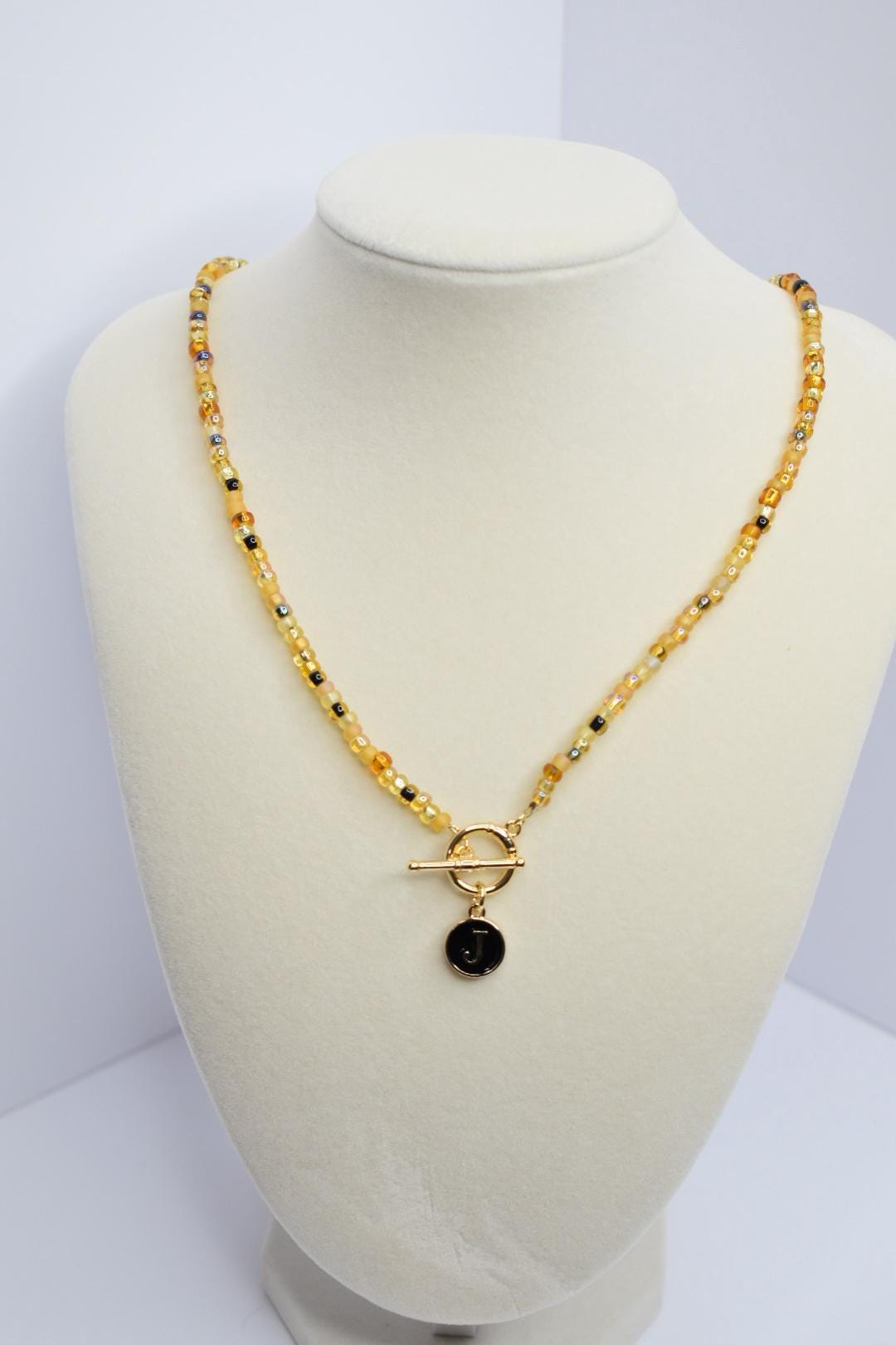A beaded necklace with a gold toggle clasp and a small, round pendant. The necklace features small, round beads in shades of yellow, amber, and black. The pendant is black with a gold-colored letter 'J' on it. The necklace is displayed on a white bust.