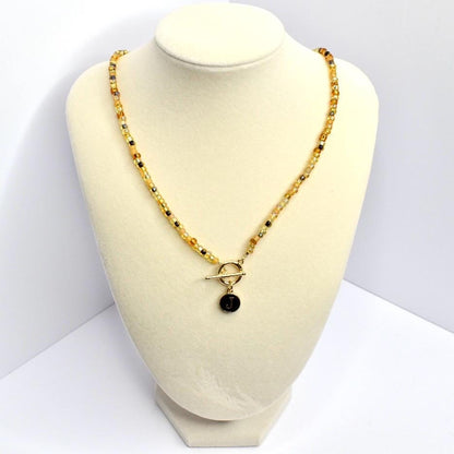 A beaded necklace with a gold-colored toggle clasp and a pendant. The necklace features small, round beads in shades of yellow, gold, and black. The pendant is a small, black circle with the letter 'J' in gold. The necklace is displayed on a cream-colored bust.