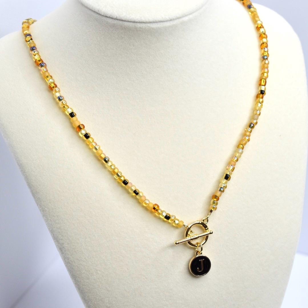 A beaded necklace with a toggle clasp and a pendant. The necklace features small, round beads in shades of yellow, gold, and black. The toggle clasp is gold-colored, and the pendant is a small, black circle with the letter 'J' in white.