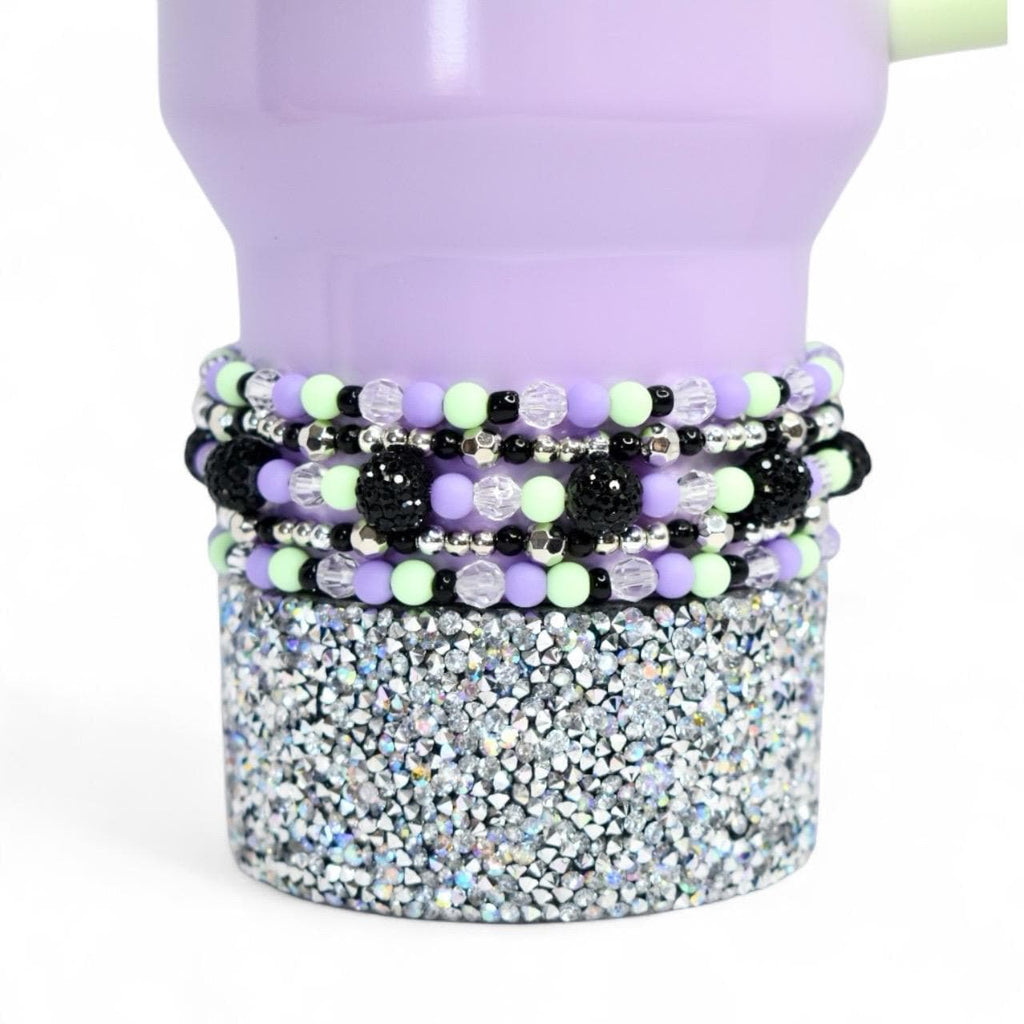 A lavender tumbler with a silver glitter base and beaded bracelets in black, silver, light green, and lavender. The bracelets are adorned with crystal and black beaded accents. The tumbler has a light green straw.