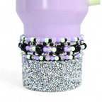 A lavender tumbler with a silver glitter base and beaded bracelets in black, silver, light green, and lavender. The bracelets are adorned with crystal and black beaded accents. The tumbler has a light green straw.
