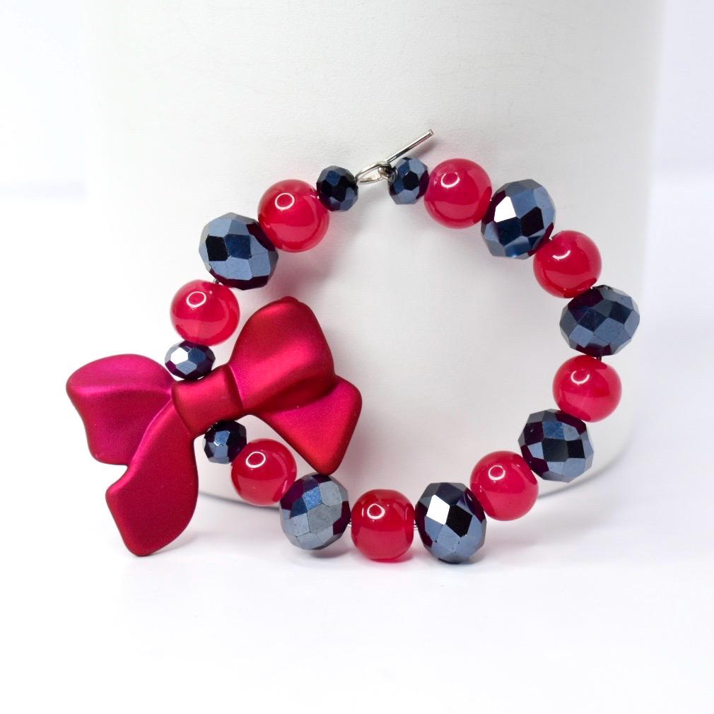 A bracelet featuring a large, fuchsia bow and a series of red and dark gray beads. The bracelet is strung together with a silver clasp. The beads are round and faceted.