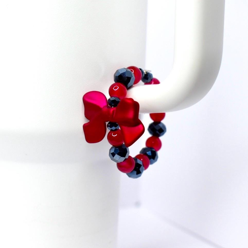 A red bow-shaped charm and a beaded bracelet with red and black beads are attached to a white mug handle. The bow is a vibrant red, and the beads are a mix of red and faceted black.