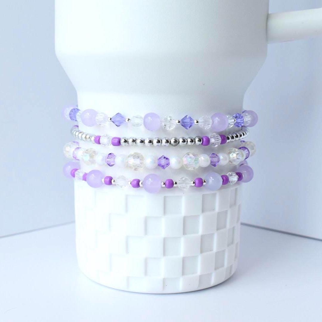 Stack of beaded bracelets in shades of lavender, purple, and clear. The bracelets are made with various beads, including round and faceted beads, and are displayed on a white, textured surface.