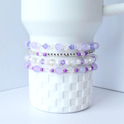 Stack of beaded bracelets in shades of lavender, purple, and clear. The bracelets are made with various beads, including round and faceted beads, and are displayed on a white, textured surface.