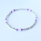 A delicate beaded bracelet featuring small silver beads interspersed with clear and purple beads. The bracelet is circular and sits on a white surface. The jewelry is simple and elegant.