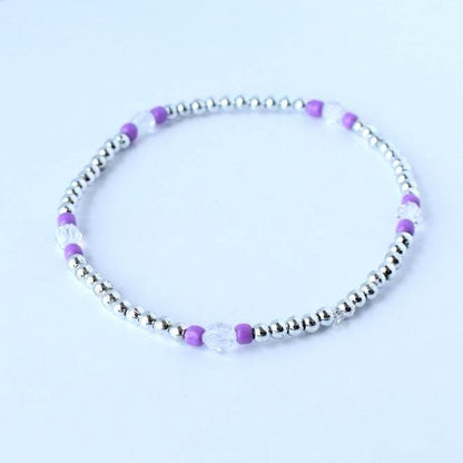 A delicate beaded bracelet featuring small silver beads interspersed with clear and purple beads. The bracelet is circular and sits on a white surface. The jewelry is simple and elegant.