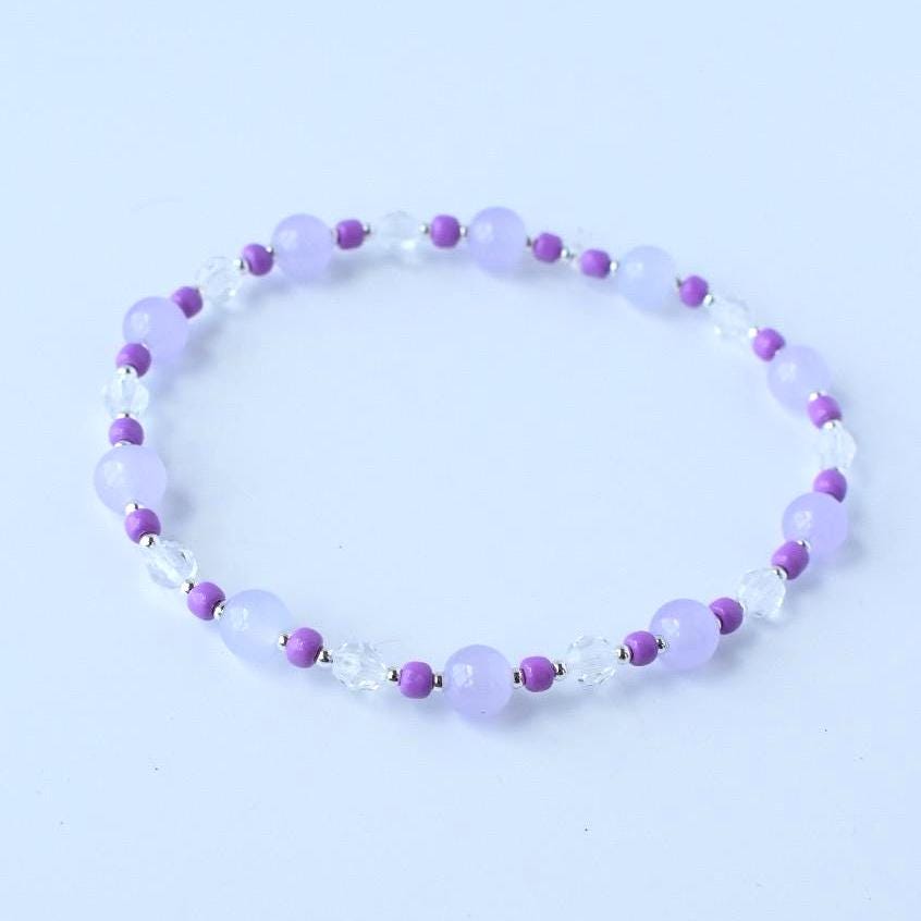A delicate bracelet featuring alternating lavender and clear beads. The lavender beads are round and smooth, while the clear beads are faceted. Small, dark purple beads separate the larger beads, creating a subtle contrast. The bracelet is designed to be worn around the wrist.