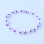 A delicate bracelet featuring alternating lavender and clear beads. The lavender beads are round and smooth, while the clear beads are faceted. Small, dark purple beads separate the larger beads, creating a subtle contrast. The bracelet is designed to be worn around the wrist.