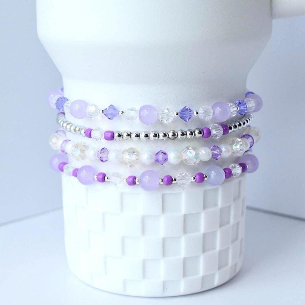 Stack of four beaded bracelets in shades of purple, white, and silver. The bracelets are made with various bead sizes and shapes, including round, faceted, and crystal beads. The bracelets are displayed on a white ceramic object.