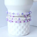 Stack of four beaded bracelets in shades of purple, white, and silver. The bracelets are made with various bead sizes and shapes, including round, faceted, and crystal beads. The bracelets are displayed on a white ceramic object.