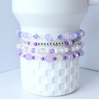 Stack of four beaded bracelets in shades of purple, white, and silver. The bracelets are made with various bead sizes and shapes, including round, faceted, and crystal beads. The bracelets are displayed on a white ceramic object.