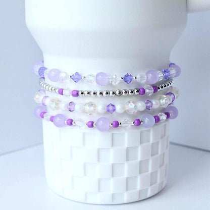 Stack of four beaded bracelets in shades of purple, white, and silver. The bracelets are made with various bead sizes and shapes, including round, faceted, and crystal beads. The bracelets are displayed on a white ceramic object.