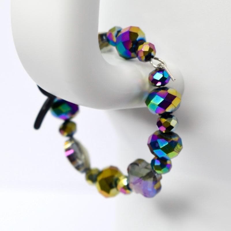 A tumbler handle bracelet with iridescent beads. The beads are various sizes and shapes, including round and faceted. The colors shift between blues, greens, purples, and yellows. The bracelet is strung on a silver chain and has a clasp closure. The beads are clustered together.