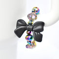 A black bow-shaped charm is the focal point of a tumbler handle bracelet. The charm is connected to a strand of iridescent, faceted beads that reflect multiple colors. The beads are spherical and add a touch of sparkle to the jewelry. The bracelet is displayed against a white background.