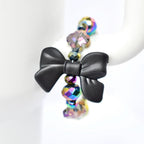 A black bow-shaped charm is the focal point of a tumbler handle bracelet. The charm is connected to a strand of iridescent, faceted beads that reflect multiple colors. The beads are spherical and add a touch of sparkle to the jewelry. The bracelet is displayed against a white background.