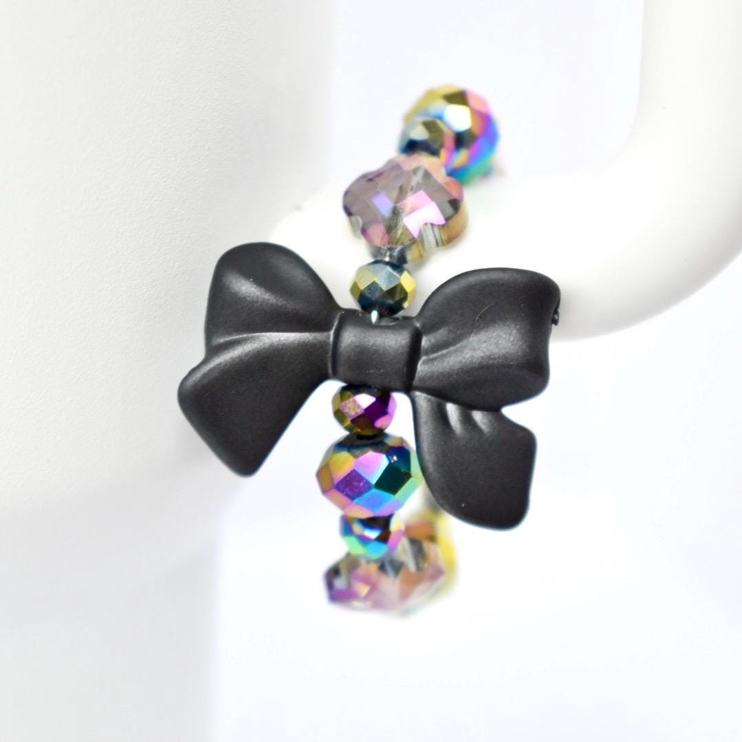 A black bow-shaped charm is the focal point of a tumbler handle bracelet. The charm is connected to a strand of iridescent, faceted beads that reflect multiple colors. The beads are spherical and add a touch of sparkle to the jewelry. The bracelet is displayed against a white background.