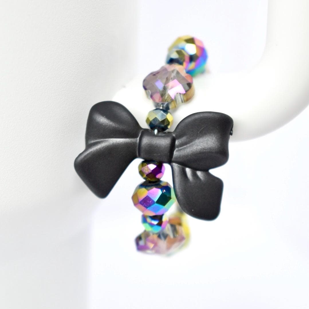 A black bow-shaped charm is the focal point of a tumbler handle bracelet. The charm is connected to a strand of iridescent, faceted beads that reflect multiple colors. The beads are spherical and add a touch of sparkle to the jewelry. The bracelet is displayed against a white background.