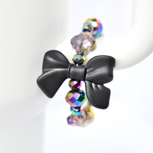 A black bow-shaped charm is the focal point of a tumbler handle bracelet. The charm is connected to a strand of iridescent, faceted beads that reflect multiple colors. The beads are spherical and add a touch of sparkle to the jewelry. The bracelet is displayed against a white background.